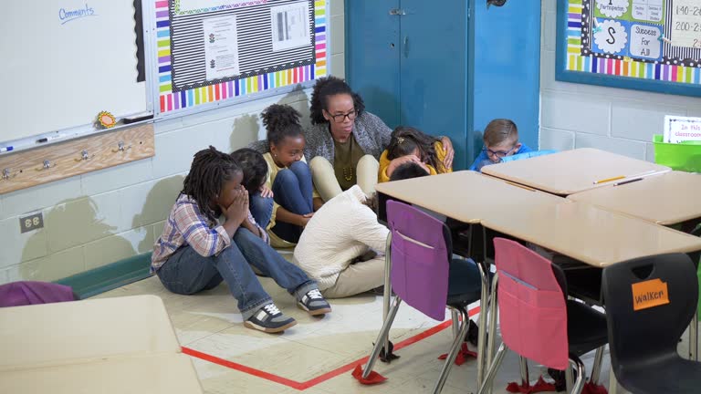 A multiracial group of seven school children and their teacher doing a practice drill, sheltering in place. They are huddled together, sitting on the floor in a corner of their classroom.